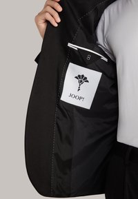 Schwarze Jacke mit glatter Textur, ausgestattet mit einem weißen Etikett mit einem Logo und einer Nummer sowie einem eleganten Innenfutter. Akzente umfassen sichtbare Nähte.