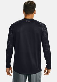 Mężczyzna widziany z tyłu, ubrany w czarną, długą koszulkę sportową z małym logo na górnej części pleców oraz szare szorty.