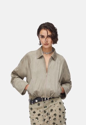 Donna con capelli corti e ricci, indossa una giacca beige con cerniera e una gonna beige decorata con paillettes, in piedi con le mani nelle tasche su sfondo bianco.