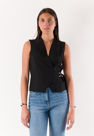 Donna che indossa un gilet nero senza maniche con fibbia laterale e jeans blu, in piedi su sfondo bianco semplice.