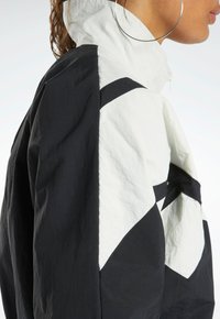 Mustvalge jakk kõrge kaelusega, millel on geomeetrilised mustrid, lukuga sulgemine ja kerge tekstuuriga kangas.