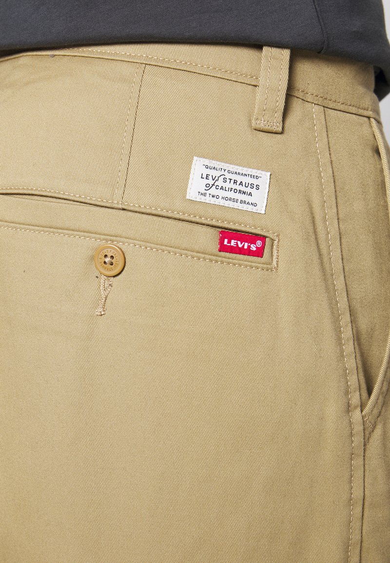Pantaloni khaki in cotone con una superficie liscia. Presentano un'etichetta del marchio Levi's e un bottone marrone. La cucitura è visibile lungo le cuciture.