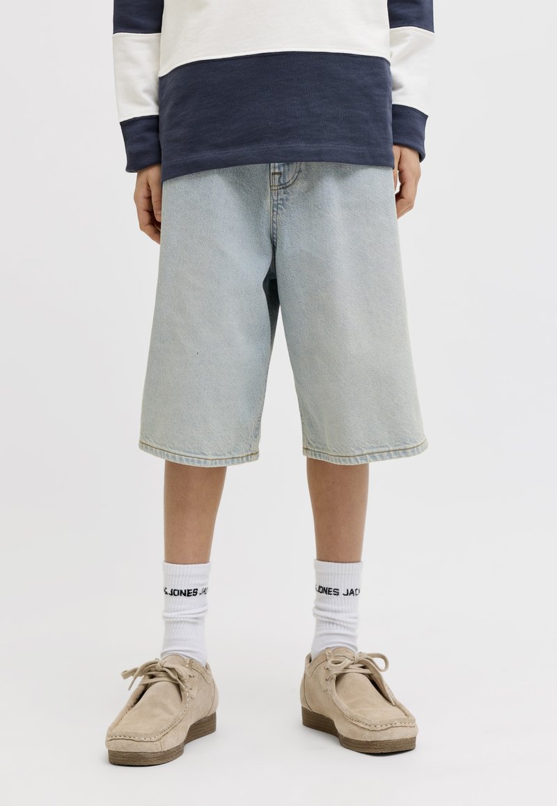 Persona che indossa pantaloncini lunghi in denim azzurro chiaro, calzini bianchi con marchio nero, mocassini beige e un maglione a righe blu navy e bianco.