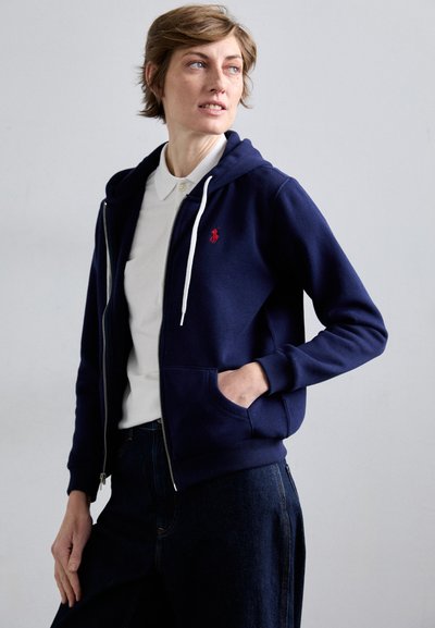 Marineblaue Kapuzenjacke aus weichem Material mit einem roten Logo auf der linken Brust, weißen Kordeln und Fronttaschen, kombiniert mit einem weißen Hemd mit Kragen.