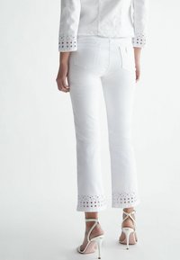 Pantalones de denim blanco con dobladillo acampanado que presentan detalles de ojales. Combinados con sandalias de tacón alto de color claro. Textura suave.
