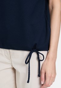 Granatowa, teksturowana bluzka z wiązaniem na dole, zestawiona z jasnobeżowymi spodniami. Prezentuje wzór tkaniny plecionej i swobodny design.