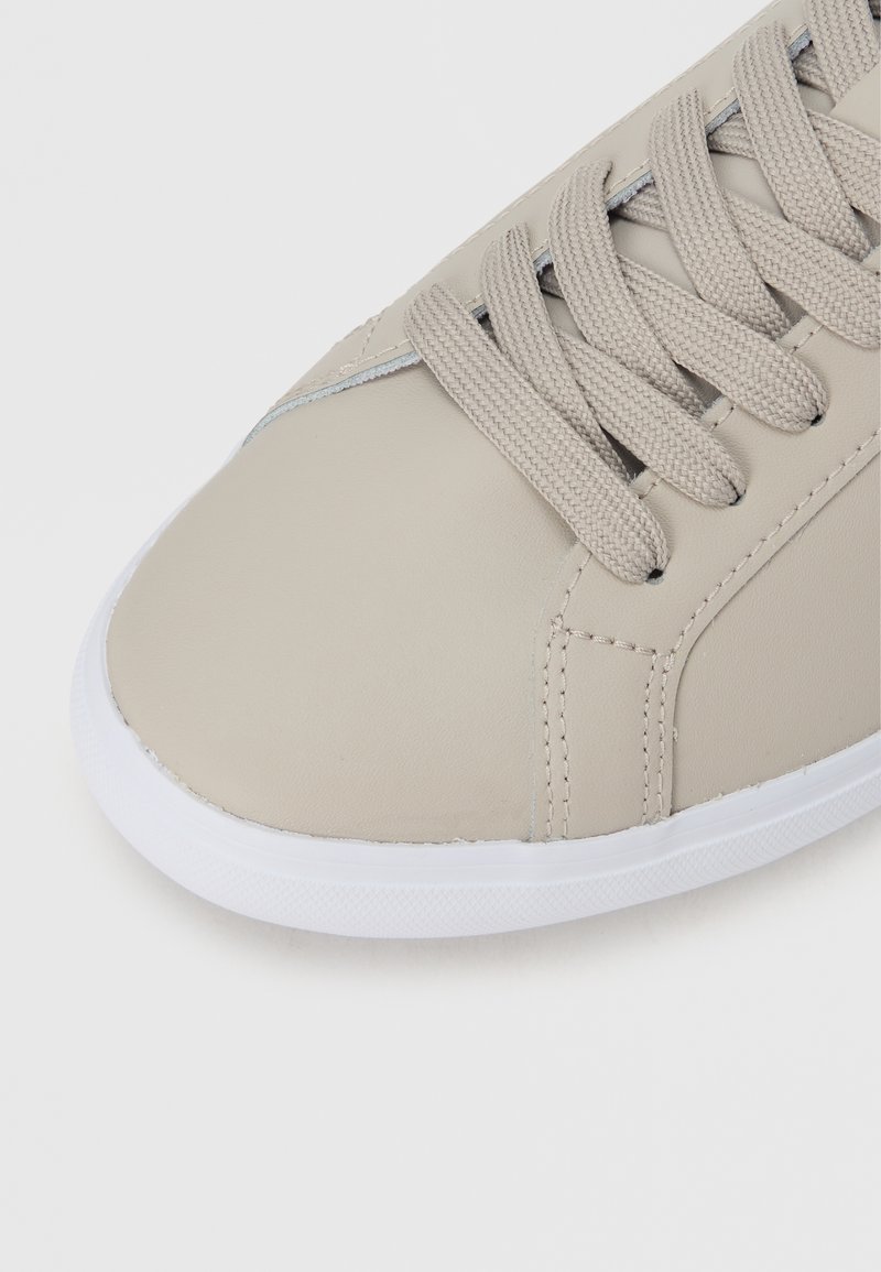 Beige sneaker med en glat syntetisk overdel, rund tå og flad hvid gummisål. Har flade snørebånd og minimale syninger.