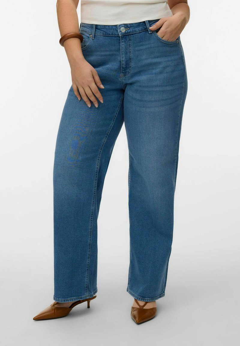 Femme portant un jean bleu taille haute à jambes droites, un haut blanc rentré, des talons marron à bout pointu et un bracelet marron au poignet gauche.