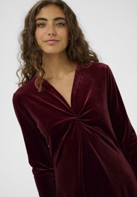 Burgundi bársonydressz hosszú ujjakkal, mély V-nyakkal és középen csavart részlettel a mellrészen; sima textúra és testhezálló fazon.