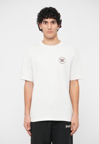 Λευκό βαμβακερό μπλουζάκι με στρογγυλή λαιμόκοψη και κοντά μανίκια. Διαθέτει ένα μικρό καφέ λογότυπο εκτυπωμένο στο αριστερό στήθος.