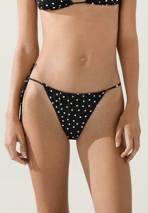 Parte inferiore del bikini nera con pois bianchi legata ai lati sulla vita e sui fianchi di una donna su sfondo neutro.