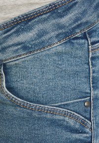 Blå denim tyg med en framficka med en metallnitar, synlig söm och en lätt sliten textur.