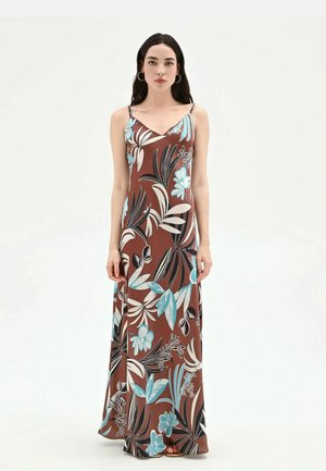 Donna in piedi con un abito lungo marrone senza maniche a motivi floreali tropicali blu, bianchi e neri, rivolta in avanti davanti a uno sfondo semplice.