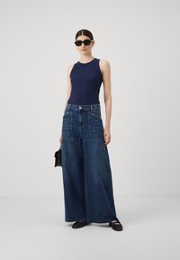 Mornarsko plava majica bez rukava u kombinaciji s širokim tamnoplavim trapericama koje imaju velike prednje džepove i sirovi rub. Dovršena s crnim dodacima.