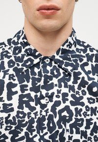 Άνδρας που φοράει πουκάμισο με κουμπιά με ασπρόμαυρο αφηρημένο μοτίβο γραμμάτων και χρυσούς κρίκους στα αυτιά.