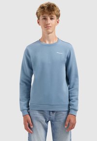 Tienerjongen in een lichtblauwe sweatshirt met ronde hals en de tekst "BALLIN'" erop, met een lichte spijkerbroek, staand tegen een effen achtergrond.
