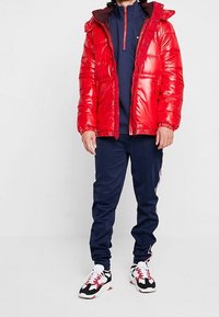 Röd puffjacka med glänsande yta och huva, marinblå zip-up-tröja, marinblå träningsbyxor med sidostråk och mönstrade sportskor.