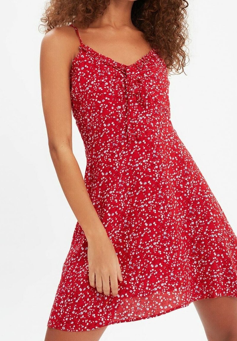 Femme portant une robe rouge sans manches avec un petit imprimé floral blanc et bleu, debout devant un fond clair uni.