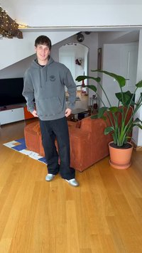 Γκρι φούτερ με κουκούλα και τσέπη μπροστά, φαρδιά μαύρη παντελόνια και ασημένια παπούτσια. Εσωτερικός χώρος με γλάστρα και πορτοκαλί καναπέ.