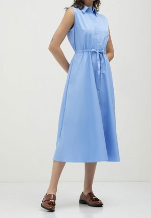 Bezrekawna, jasnoniebieska sukienka midi z kołnierzykiem i paskiem w talii, noszona przez stojącą osobę, uzupełniona brązowymi skórzanymi mokasynami z frędzlami.