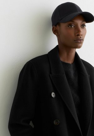 Sieviete, kas valkā melnu mēteļu, melnu džemperi un melnu cepuri, stāv pret baltu teksturētu sienu, skatoties nedaudz uz sāniem.