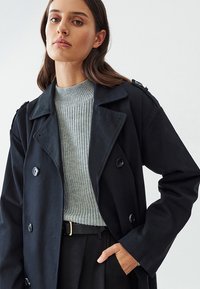 Mustana kahekordne trench coat õlavarre epaulettidega halli ribakudumiga sviitri peal. Omab suuri nuppe ja vabalt langevat lõiget. Matte tekstuur.