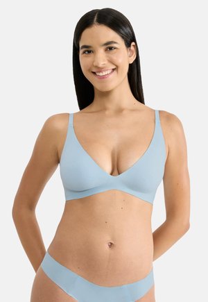 Uśmiechnięta kobieta o długich ciemnych włosach, ubrana w jasnoniebieski bezprzewodowy biustonosz i dopasowane majtki, stojąca z rękami za plecami.