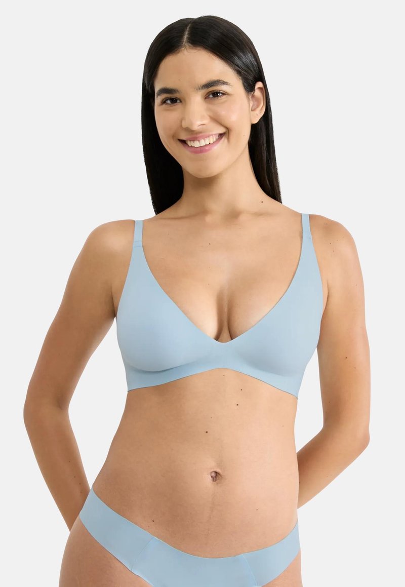 Γυναίκα με μακριά σκούρα μαλλιά που χαμογελά, φοράει ανοιχτό μπλε ασύρματο σουτιέν και ταιριαστά εσώρουχα, στέκεται με τα χέρια πίσω από την πλάτη.