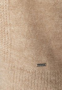 Béžový pletený svetr s texturovaným povrchem, žebrovaným lemem a kovovou štítkovou značkou. Látka působí měkce a teple, představující světlý, přírodní odstín.