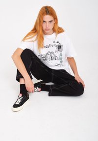 Witte t-shirt met zwarte grafische print, gecombineerd met zwarte wijdvallende broek met contrasterende stiksels, en zwarte sneakers met witte zool.