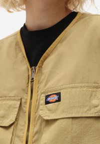 Nära bild av en tan zip-up jacka med fickor med lock och en svart skjorta under.
