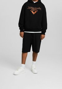 Felpa nera con grafica arancione, abbinata a pantaloncini neri. Scarpe da ginnastica bianche e calzini completano l'outfit. Stile sportivo casual.