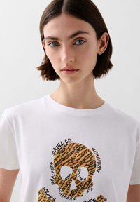 Witte katoenen t-shirt met een grafische print van een schedel in oranje en zwarte tijgerstrepen, omringd door tekstelementen.
