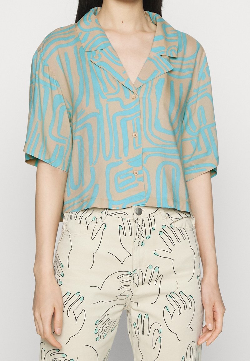 Femme portant une chemise beige à manches courtes avec des lignes abstraites bleues et un pantalon beige avec des motifs de mains noirs et turquoise.