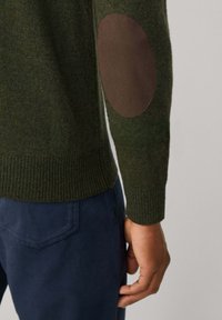 Persoon in een groene gebreide trui met een bruine elleboogpatch en donkere blauwe broek, met een gedeeltelijke arm en hand zichtbaar tegen een neutrale achtergrond.