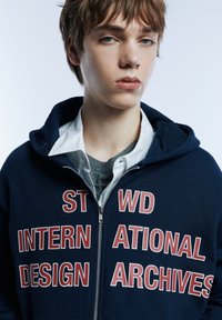 Marineblauer Kapuzenpullover mit Reißverschluss und markanten roten Buchstaben mit den Worten "INTERNATIONAL DESIGN ARCHIVES." Darüber ein gestreiftes Hemd getragen.