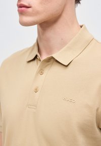 Gros plan sur une personne portant un polo beige avec un col, deux boutons et un discret logo "HUGO" sur la poitrine.