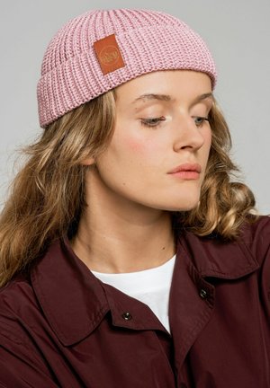 Różowa dzianinowa czapka typu beanie z prążkowaną fakturą i brązową skórzaną metką. Noszona z ciemną kurtką, prezentująca zbliżenie na szwy i detale wzornicze.