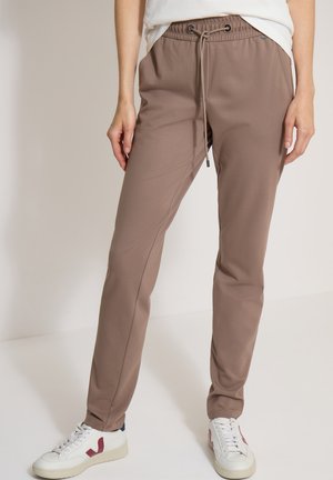 Personne portant un pantalon taupe avec cordon et des baskets blanches avec des accents rouges, debout devant un fond clair uni.