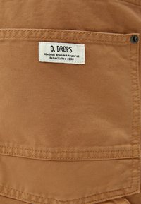 Beżowa bawełniana tkanina z naszytą metką z napisem "O. DROPS Minimal Branded Apparel Established 1998." Widoczne detale szwów.