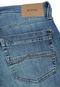 Jeans de denim azul con una etiqueta de cuero marrón que dice "MUSTANG", diseño de cinco bolsillos y costuras en contraste en los bolsillos traseros.