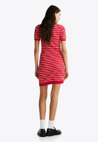 Rotes Strickkleid mit horizontalem weißen Text, der wiederholt "tommy" und "years" zeigt. Kurze Ärmel, figurbetonte Passform, knielange Länge, kombiniert mit schwarzen Schuhen.