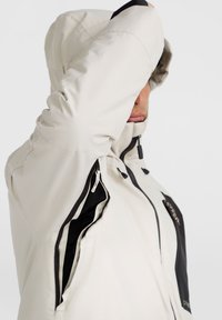 Veste à capuche beige clair avec un design accent noir. Elle dispose de poches latérales zippées et d'un tissu texturé. Capuche ajustable avec cordons.
