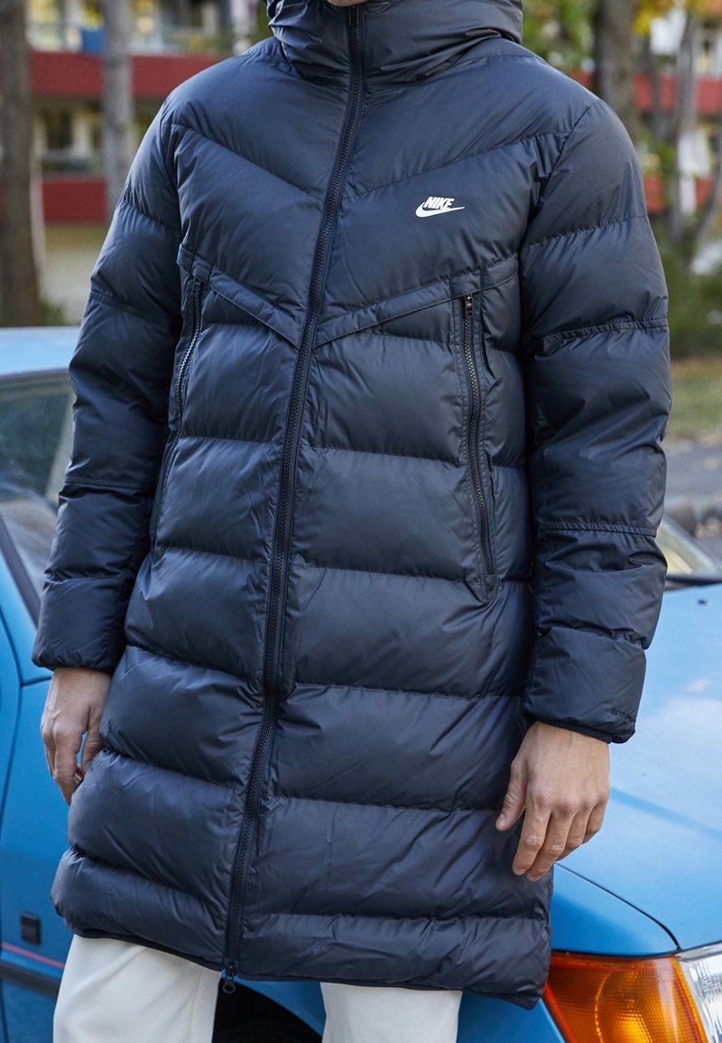 Marinblå puffjacka med huva, utrustad med fickor med dragkedja, ett quiltat mönster och en liten vit logotyp på övre vänstra sidan.