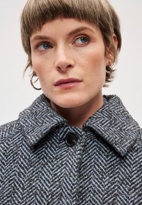 Pelēkas herringbone stila mētelis ar plašu apkakli, lielu melnu pogu un raksturīgu audumu. Modelim ir īsi mati un gredzenveida auskari.