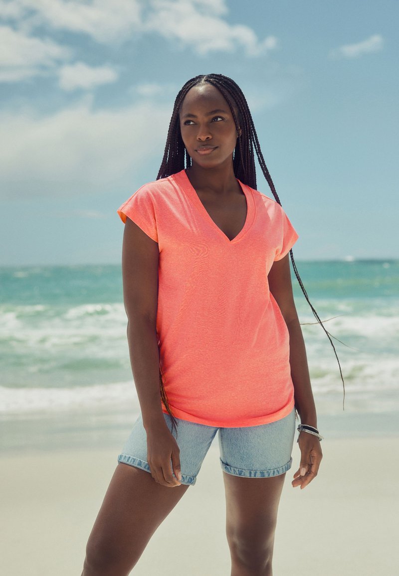 Sieviete ar gariem bizēm, kas valkā korāla krāsas V-veida izgriezuma kreklu un džinsa šortus, stāv saulainā pludmalē ar okeāna viļņiem fonā.