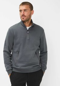 Felpa grigia con zip a quarti, dal texture morbido, presenta due tasche laterali con zip e un orlo a costine. Indossata sopra una camicia bianca.