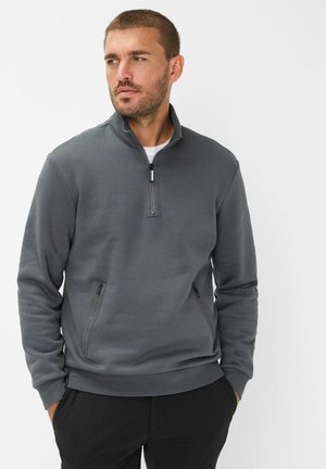 Felpa grigia con zip a quarti, dal texture morbido, presenta due tasche laterali con zip e un orlo a costine. Indossata sopra una camicia bianca.