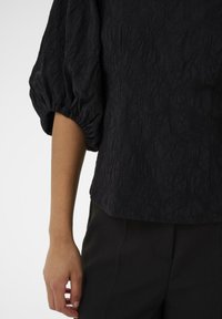 Melnas teksturētas blūzes ar puffed piedurknēm, kurai ir sarautas zīmējums un brīva piegriezuma. Apvienota ar piegrieztiem melniem biksēm.