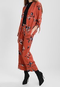 Oranžový kostým s květinovým potiskem z lehkého materiálu, zahrnující sako s ohrnutými rukávy a široké nohavice. Stylizováno s černými botami na podpatku.